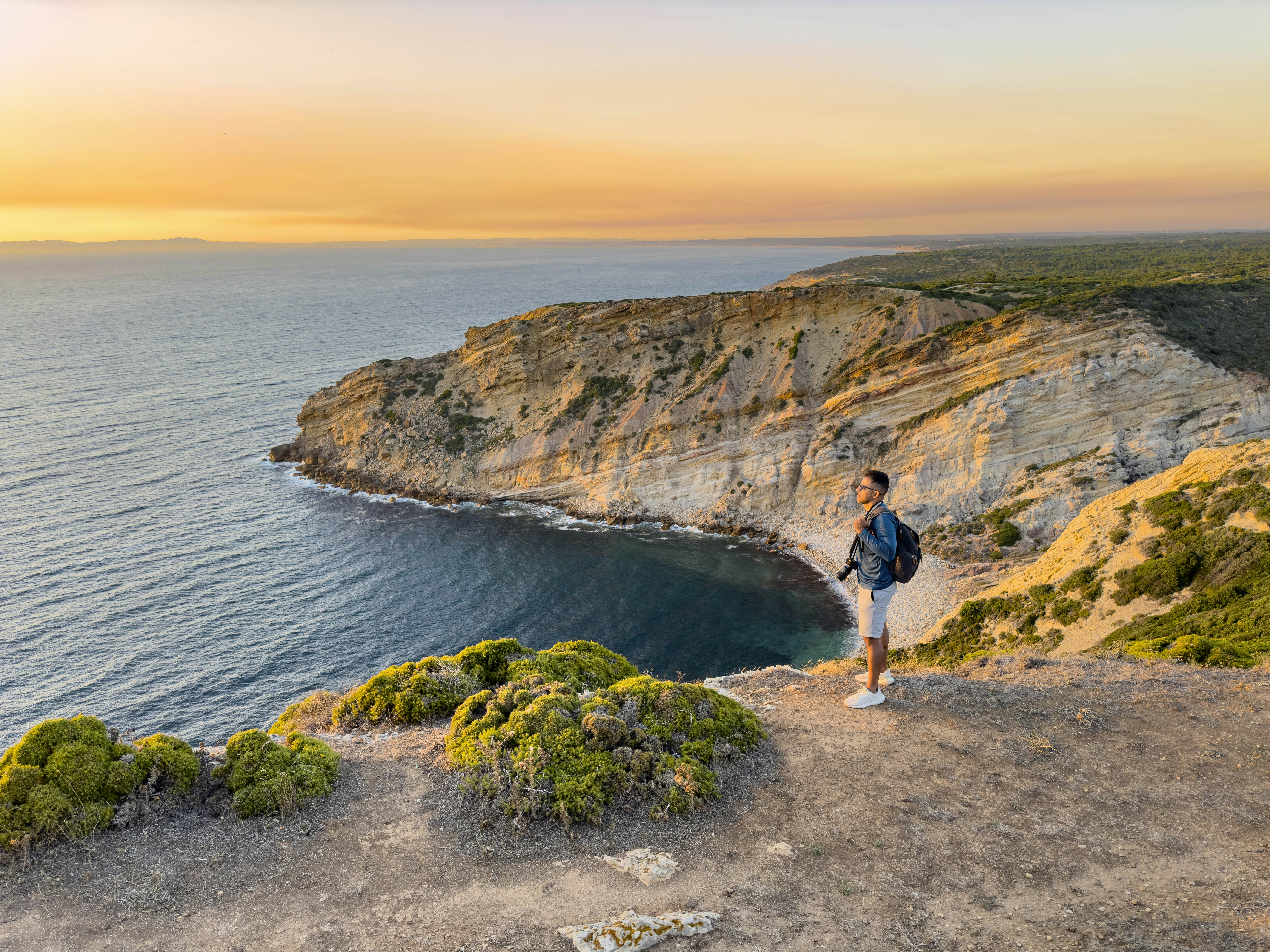 A person with a camera overlooks a stunning sunset from a cliffside beach on the Setúbal Peninsula, Portugal.