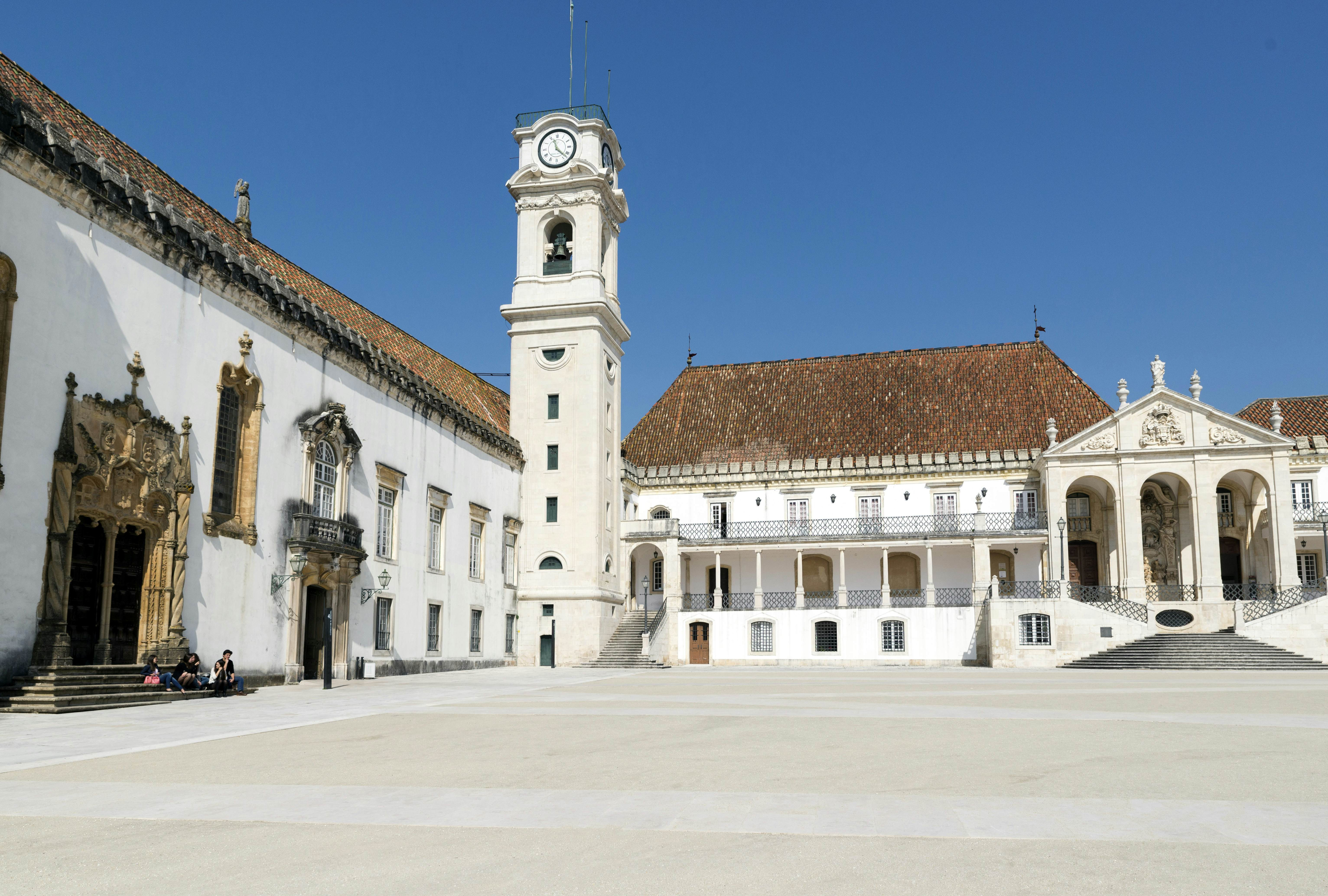 A wide city square in Coimbra, Portugal, showcases historic buildings and a prominent clock tower against a clear sky.