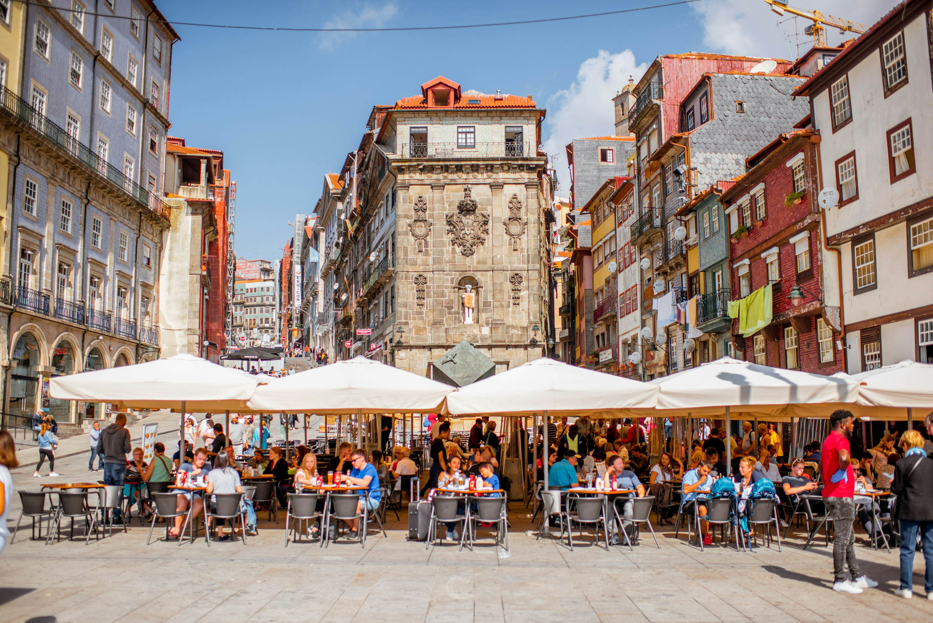 A bustling city square in Porto, Portugal, is filled with people enjoying cafes under umbrellas in the Ribeira district.