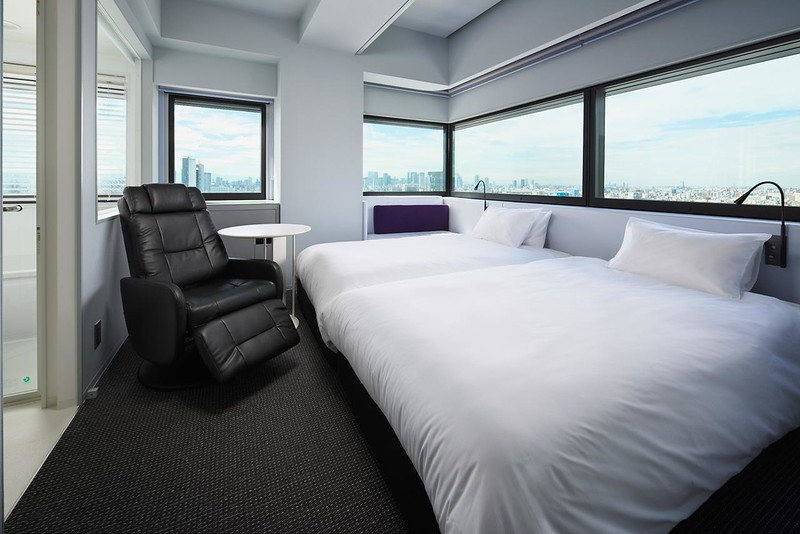 Twin guest room in the Remm Shin-Osaka hotel, Osaka, Japan, with two large beds, pushed together, beneath corner windows overlooking the city during the day, and leather comfort chair