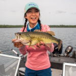 Woman smiling wearing a salmon colored hooded fishing shirt and sunglasses, holding a fishing rod.