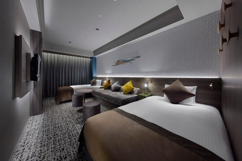 Compact twin guest room in the Hotel Villa Fontaine Grand Tokyo-Roppongi, Japan, with two beds, separated by a three-man sofa, opposite a wall-mounted TV, and pleated curtain drawn against the window